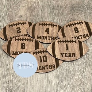May include: Wooden football-shaped milestone markers with laser-etched details. Each marker displays a number indicating months or a year, ranging from 2 to 10 months and 1 year. The Branding Spot logo is visible.