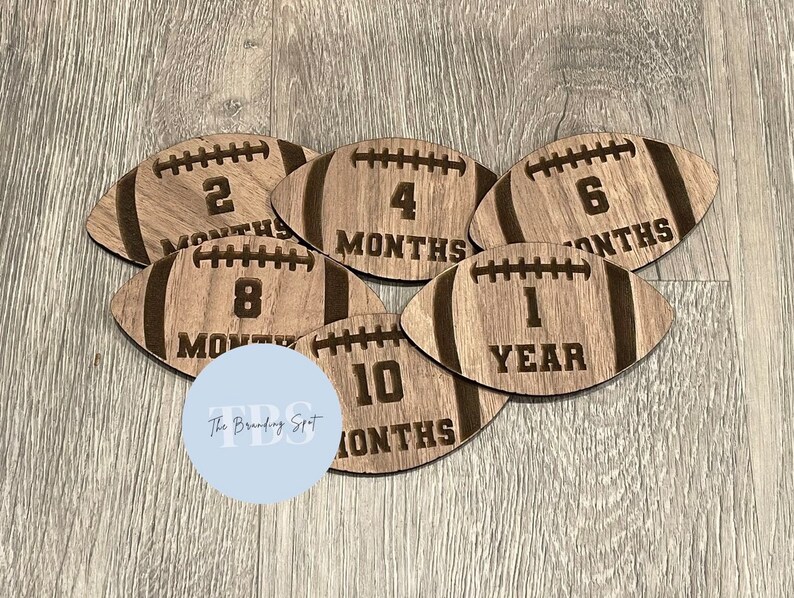 May include: Wooden football-shaped milestone markers with laser-etched details. Each marker displays a number indicating months or a year, ranging from 2 to 10 months and 1 year. The Branding Spot logo is visible.