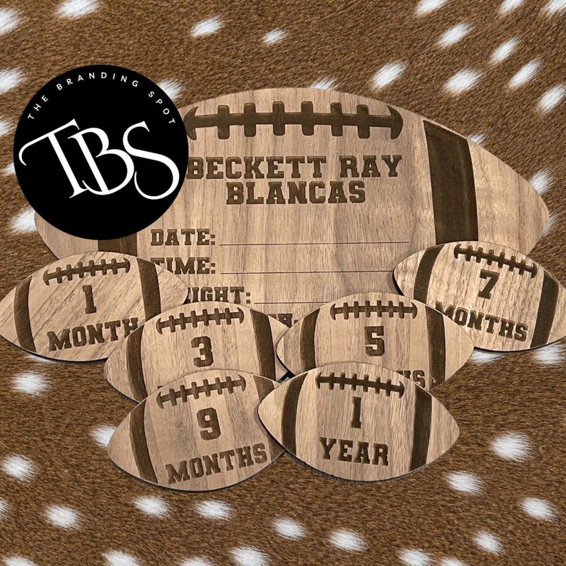 May include: A wooden football-shaped milestone board with the name "BECKETT RAY BLANCAS" engraved. Includes smaller footballs marked 1, 3, 5, 7, and 9 months, and 1 year. The board has spaces for date, time, and height.