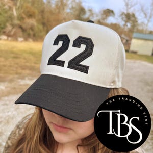 May include: A baseball cap with a white crown and a black brim. The number "22" is embroidered in black on the front. The cap has a curved brim and a black button on top. The logo "The Branding Spot" is on the bottom right.