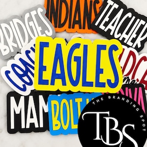 May include: A collection of colorful, layered silicone-style name and word tags featuring various text including EAGLES, INDIANS, TEACHER, BRIDGES, COACH, BOLT, and MAM. The tags are displayed in a stacked arrangement with bold, high-contrast lettering.
