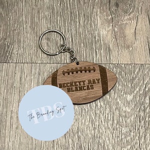 May include: A wooden football-shaped keychain with the name "BECKETT RAY BLANCAS" engraved on it. The keychain has a silver metal ring and chain. The football has a light brown color with darker brown details.