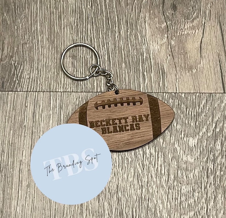 May include: A wooden football-shaped keychain with the name "BECKETT RAY BLANCAS" engraved on it. The keychain has a silver metal ring and chain. The football has a light brown color with darker brown details.