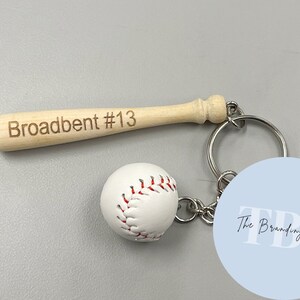 May include: A wooden baseball bat keychain with the name "Broadbent #13" engraved on it. The keychain also features a white baseball and a blue circle with the text "The Branding Spot" in white.