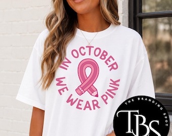 May include: A white short-sleeved t-shirt featuring a pink awareness ribbon shaped like a pencil in the center. The text surrounding the ribbon reads IN OCTOBER WE WEAR PINK in a circular pink font. The design is printed on the front of the shirt.