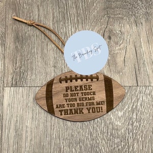 May include: A wooden football-shaped sign with the text "PLEASE DO NOT TOUCH YOUR GERMS ARE TOO BIG FOR ME! THANK YOU!". It has a leather cord for hanging. The sign is light brown with dark brown engraved text.
