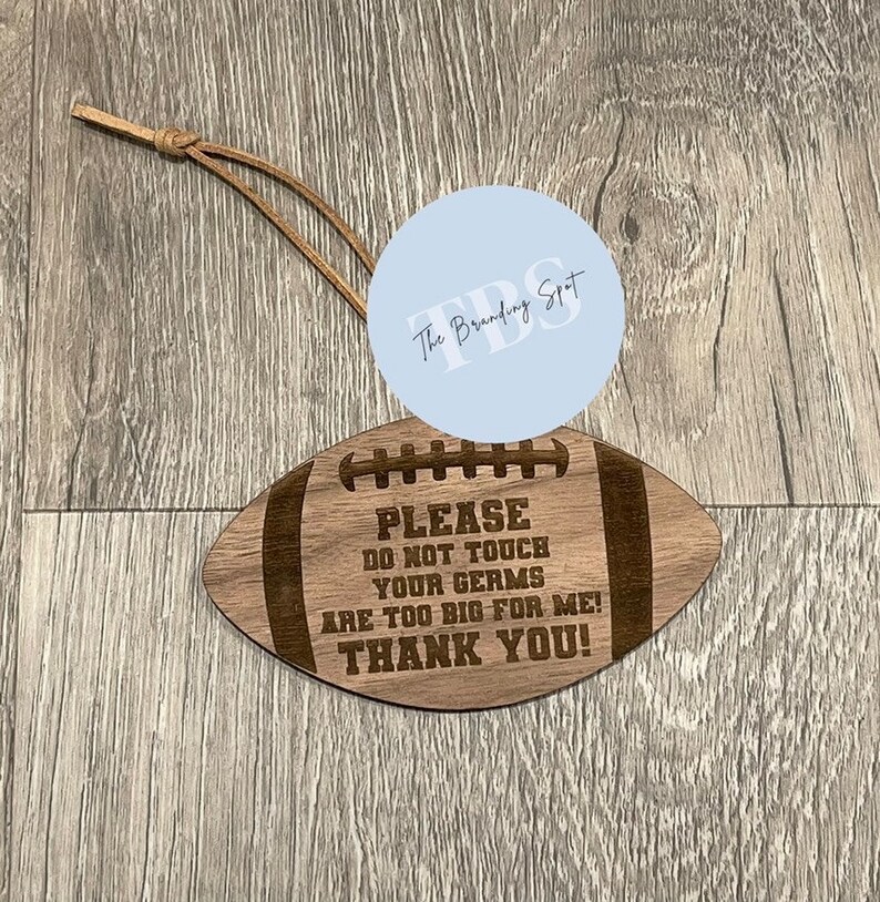 May include: A wooden football-shaped sign with the text "PLEASE DO NOT TOUCH YOUR GERMS ARE TOO BIG FOR ME! THANK YOU!". It has a leather cord for hanging. The sign is light brown with dark brown engraved text.