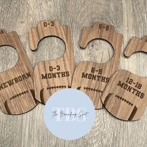 May include: Four wooden baby clothes hangers with football designs. Each hanger is labeled with a different size: Newborn, 0-3 Months, 6-9 Months, and 12-18 Months. The hangers are made of light brown wood.