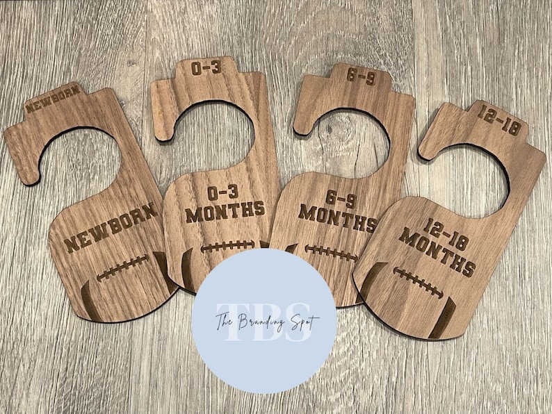 May include: Four wooden baby clothes hangers with football designs. Each hanger is labeled with a different size: Newborn, 0-3 Months, 6-9 Months, and 12-18 Months. The hangers are made of light brown wood.