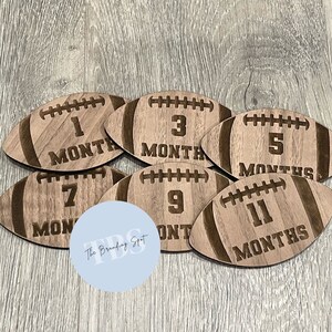 May include: A set of six wooden football-shaped monthly milestone markers. Each marker is engraved with a number and the word "MONTHS", indicating the age of a baby. The markers are made of light brown wood.