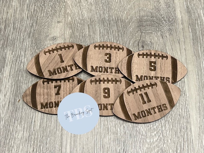 May include: A set of six wooden football-shaped monthly milestone markers. Each marker is engraved with a number and the word "MONTHS", indicating the age of a baby. The markers are made of light brown wood.