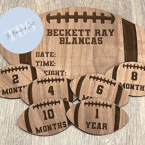 May include: A wooden football-shaped milestone sign with the name "BECKETT RAY BLANCAS" engraved at the top. Includes smaller footballs marked "2 MONTHS", "4", "6 MONTHS", "8 MONTHS", "10 MONTHS", and "1 YEAR".