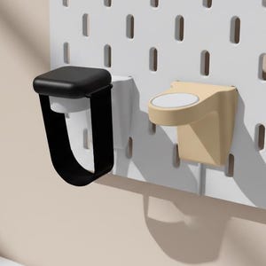 Könnte beinhalten: Zwei Wandhalterungen für Smartwatches, eine schwarze und eine beige. Der schwarze Halter hat einen U-förmigen Riemen. Der beige Halter ist kreisförmig. Beide sind an einer weißen Lochwand befestigt. Eine beige Vase mit Grün steht auf einer weißen Oberfläche.