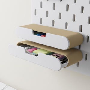 May include: Two-tiered, wall-mounted storage unit with a white and tan color scheme. The unit is attached to a white pegboard. A white keyboard, mouse, and small potted succulent are on a white desk in the foreground. The drawers contain office supplies.