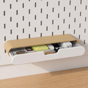 Könnte beinhalten: Eine weiße Lochwand mit einer beige-weißen Schublade, die verschiedene kleine Gegenstände enthält. Die Lochwand hat ein Gitter aus gleichmäßig verteilten, vertikalen Schlitzen. Die Schublade ist geöffnet und zeigt Gegenstände wie ein Ladegerät.