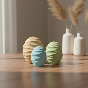 Könnte beinhalten: Drei dekorative, eiförmige Objekte in Pastellfarben: Gelb, Blau und Grün. Jedes Ei hat eine strukturierte, geriffelte Oberfläche. Zwei weiße Vasen mit getrockneten Pflanzen stehen im Hintergrund auf einem Holztisch. Die Szene wird von natürlichem Licht beleuchtet.