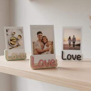 Könnte beinhalten: Drei Fotorahmen mit dem Wort "Love" an der Basis. Jeder Rahmen enthält ein anderes Foto. Die Rahmen stehen auf einem hellen Holzregal. Eine Vase mit getrockneten Pflanzen und eine Kerze befinden sich ebenfalls auf dem Regal.