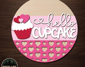 May include: A circular wooden door hanger kit featuring a cupcake graphic with three hearts on top and the text hello CUPCAKE in white script and block lettering. The lower section has a laser-cut pattern of small hearts on a pink background.