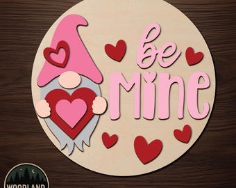 May include: A circular unfinished wooden door hanger kit featuring a gnome wearing a pink hat and holding a red heart. The text "be Mine" is written in pink script. Several red and pink hearts are arranged around the gnome and the text.