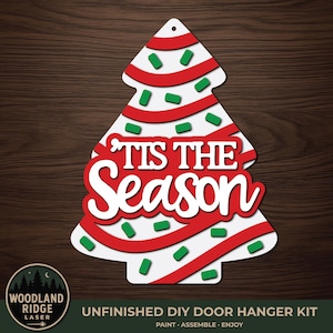 May include: A white Christmas tree-shaped door hanger kit featuring red ribbon accents and green sprinkles. The text 'TIS THE SEASON' is displayed in white and red lettering across the center. The kit includes a small hole at the top for hanging.