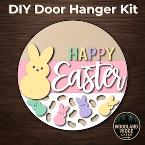May include: DIY door hanger kit with a circular wooden base. The design features a yellow bunny, pastel-colored text that reads "HAPPY Easter", and several small bunny cutouts. The background includes pink and white stripes.