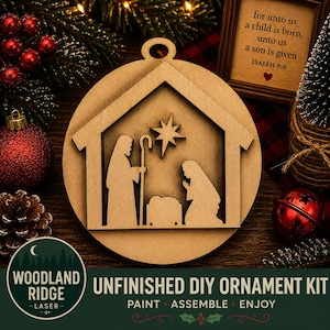 May include: A circular unfinished wood ornament kit featuring a laser-cut nativity scene with a stable, a star, and figures of two people kneeling beside a manger. The ornament has a small hole at the top for hanging and a smooth, light brown finish.