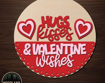 May include: A circular wooden door hanger kit featuring the text HUGS KISSES & VALENTINE WISHES in red and pink layered lettering. The design includes two heart shapes and a scalloped red border. The wood is light-coloured and unfinished.
