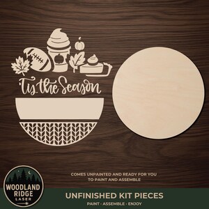May include: An unfinished laser-cut wood craft kit featuring a round base and a decorative topper with the text 'Tis the Season'. The design includes a football, a coffee cup, a pumpkin, leaves, and a slice of pie, with a knit-patterned bottom section.