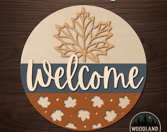May include: A circular DIY door hanger kit featuring a light wood top section with a large maple leaf cutout, a dark blue middle stripe with the word Welcome in light wood script, and a burnt orange bottom section with leaf cutouts.
