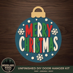 May include: A circular blue Christmas ornament door hanger kit featuring the text MERRY CHRISTMAS in red, green, and cream letters. The design includes white snowflake cutouts and small white dots. The bottom corner displays a circular logo with the text WOODLAND RIDGE LASER and the words UNFINISHED DIY DOOR HANGER KIT PAINT ASSEMBLE ENJOY.