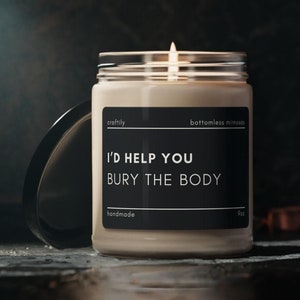 May include: A lit, cream-colored candle in a clear glass jar with a black lid. The label reads "I'd Help You Bury The Body" in white text. The candle is handmade and weighs 9 oz.