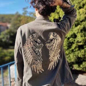 Op de afbeelding: Olijfgroene jas met gouden pailletten engelen vleugels op de rug. De jas heeft een knoopsluiting, lange mouwen en een klassieke kraag. De vleugels zijn gedetailleerd met individuele pailletten veren.