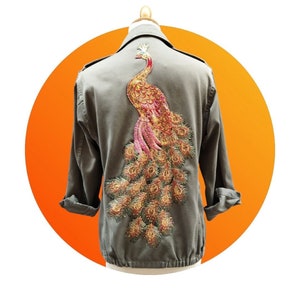 Könnte beinhalten: Olivgrüne Jacke mit einem großen, verzierten Pfau auf dem Rücken. Der Pfau hat goldene, rosa und braune Details. Die Jacke hat hochgekrempelte Ärmel und wird auf einer Schaufensterpuppe vor einem orangefarbenen Hintergrund präsentiert.