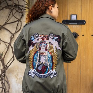 Może przedstawiać: Zielona kurtka w stylu wojskowym z dużą haftowaną naszywką na plecach. Naszywka przedstawia postać religijną z koroną, otoczoną aniołami i słowami "Tamoree Believe".