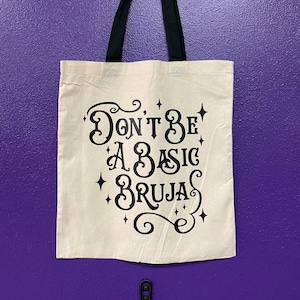 May include: A white canvas tote bag with black straps. The bag has a black design that says "Don't Be A Basic Bruja" with stars around the text.