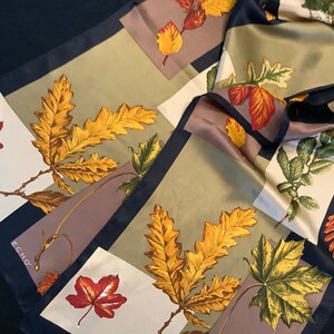 Op de afbeelding: Een zijden sjaal met een patchwork ontwerp met verschillende herfstbladeren in tinten groen, geel, rood en bruin. De sjaal heeft een zwarte rand en het woord "ECHO" is in wit gedrukt in de linkerbenedenhoek.