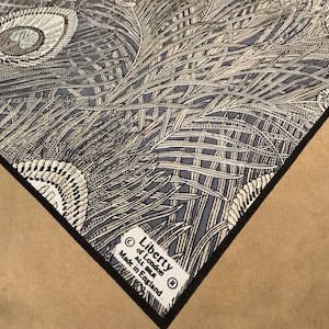 May include: A square silk scarf featuring a detailed peacock feather pattern in shades of blue, black, and white. A white fabric label is stitched to the corner with the text Liberty of London, ALL SILK, Made in England, and a black border.