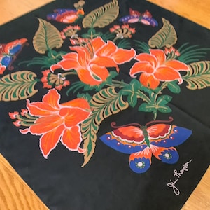 Puede incluir: Un pa&ntilde;uelo cuadrado negro con un arreglo central de lirios de color naranja brillante y follaje verde, rodeado de mariposas coloridas en tonos azules, naranjas y rojos. La firma Jim Thompson est&aacute; impresa en blanco en la esquina.