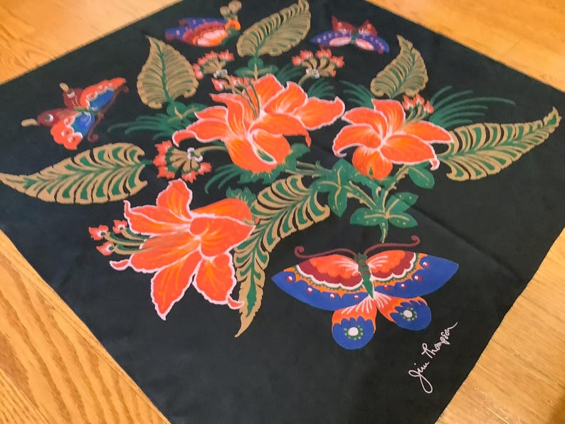 Puede incluir: Un pa&ntilde;uelo cuadrado negro con un arreglo central de lirios de color naranja brillante y follaje verde, rodeado de mariposas coloridas en tonos azules, naranjas y rojos. La firma Jim Thompson est&aacute; impresa en blanco en la esquina.