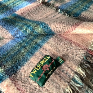 May include: A woven wool throw blanket featuring a plaid pattern in shades of soft pink, blue, and green. The blanket has a fringed edge and a green fabric label stitched onto the surface that reads Tweedmill Exclusive Product British Made.