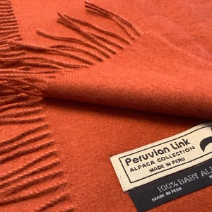 Peut inclure: Une écharpe en alpaga orange brûlé avec des bords frangés. L'écharpe est pliée, révélant une étiquette indiquant "Peruvian Link, Alpaca Collection, Made in Peru, 100% Baby Alpaca". L'écharpe est d'une couleur unie et chaude, et semble douce et luxueuse.