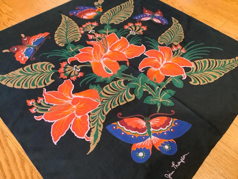 Puede incluir: Un pa&ntilde;uelo cuadrado negro con un patr&oacute;n floral central de grandes lirios naranjas, hojas verdes similares a helechos y varias mariposas coloridas en tonos azul, naranja y rojo. La firma Jim Thompson est&aacute; impresa en blanco en la esquina.