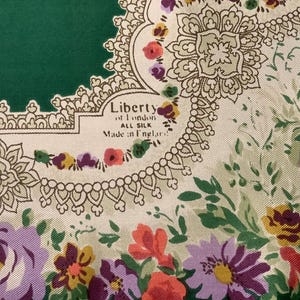 May include: A silk scarf featuring a deep green corner and a cream-colored border with intricate lace-like patterns and floral motifs in purple, red, and yellow. The text Liberty of London, ALL SILK, Made in England is printed on the fabric.