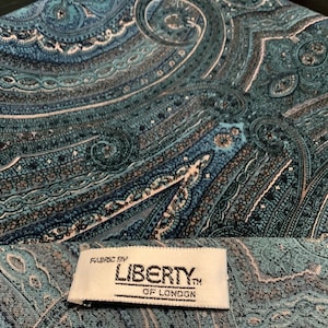 May include: Close-up of a fabric with a teal and blue paisley pattern. The fabric is labeled "Fabric by Liberty of London". The paisley design features intricate swirls and floral motifs in shades of teal, blue, and gray.