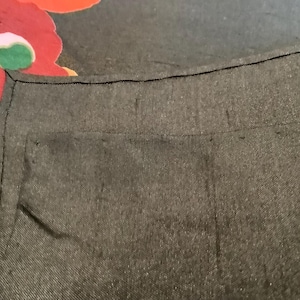 Puede incluir: Una prenda de tela gris carb&oacute;n oscuro con una costura visible a lo largo del borde superior y un detalle de bolsillo rectangular. Una parte de una tela negra con un gran estampado floral rojo, rosa y verde es visible en la esquina superior.