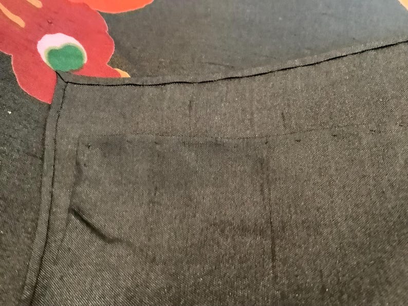 Puede incluir: Una prenda de tela gris carb&oacute;n oscuro con una costura visible a lo largo del borde superior y un detalle de bolsillo rectangular. Una parte de una tela negra con un gran estampado floral rojo, rosa y verde es visible en la esquina superior.