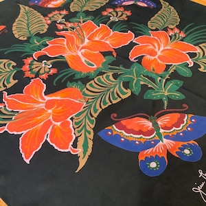 Puede incluir: Un pa&ntilde;uelo cuadrado negro con un vibrante estampado floral de grandes lirios naranjas, hojas verdes y doradas similares a helechos, y mariposas coloridas en tonos azul, naranja y rojo. La firma Jim Thompson est&aacute; impresa en blanco.