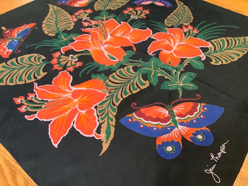Puede incluir: Un pa&ntilde;uelo cuadrado negro con un vibrante estampado floral de grandes lirios naranjas, hojas verdes y doradas similares a helechos, y mariposas coloridas en tonos azul, naranja y rojo. La firma Jim Thompson est&aacute; impresa en blanco.