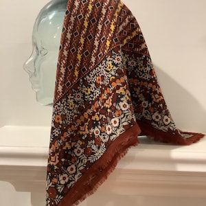 May include: A square scarf featuring a deep rust-coloured base with a multi-coloured pattern. The design includes bands of geometric diamond shapes in yellow, black, and white, alternating with sections of dense floral motifs in orange, white, and black.