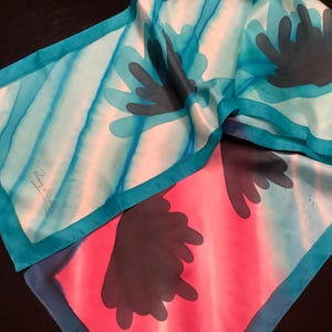 May include: A silk scarf with a vibrant design. The scarf features a teal border and a pattern of teal and dark gray floral shapes against a pink and light blue background. The scarf has a signature in the corner.
